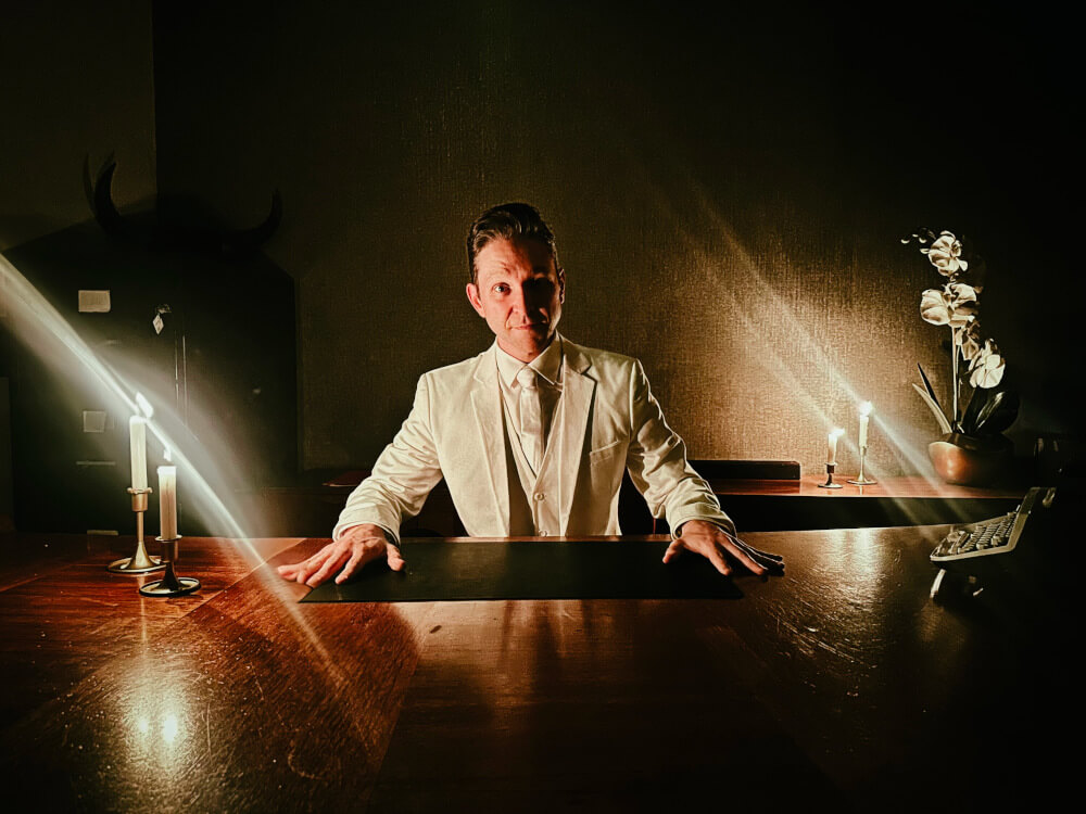 a man in a white suit sitting at a desk looking straight at you, surrounded by candles