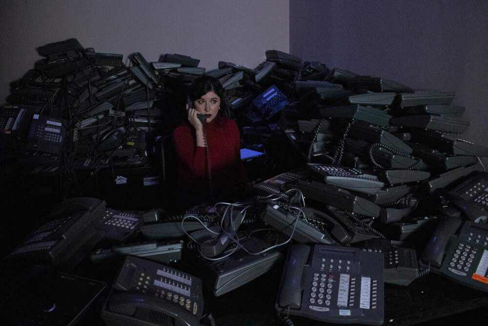 audience member in a red shirt wearing a headset sitting at a desk covered in an excessive amount of phones, listenin to one of them